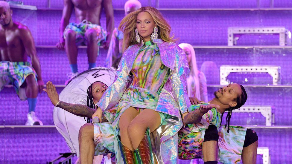 Beyonce on-stage with two dancers leaning behind her. She's dressed in a glittery-neon dress with golden boots