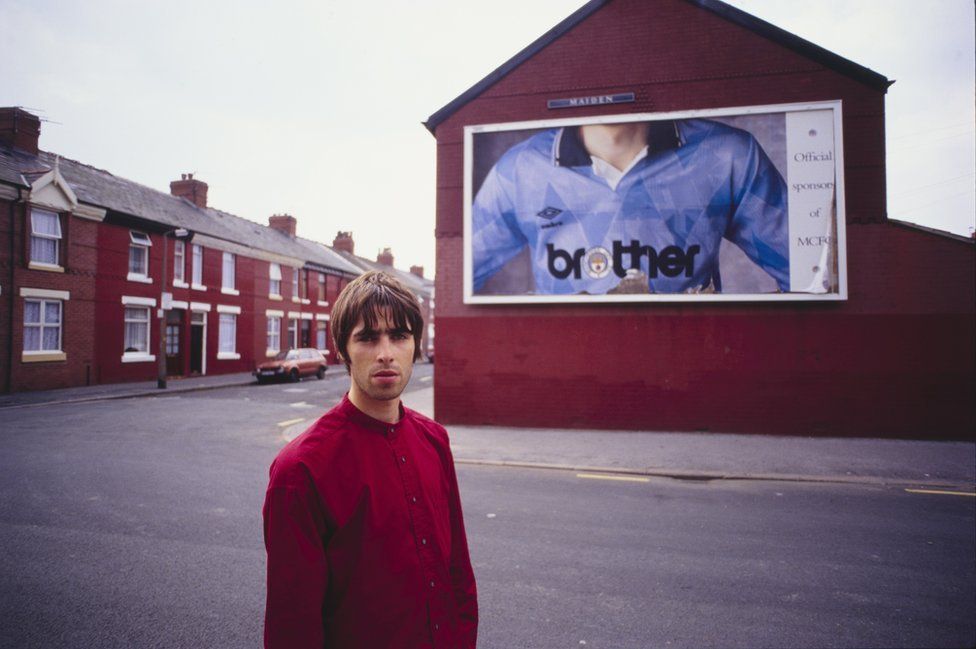Liam Gallagher stood in front of a billboard featuring a Manchester City football shirt with a "brother" logo