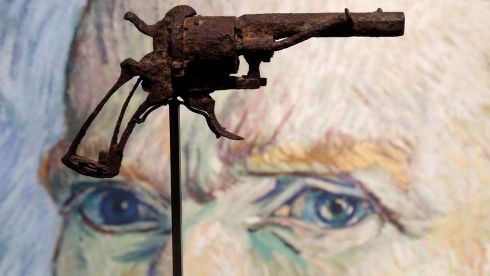 A rusted, tarnished gun raised on a display stand is seen suspended in front of a portrait of van Gogh