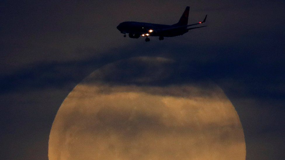 A full moon rises between clouds as a landing commercial jet approaches the airport before the start of a total lunar eclipse that is called a "Super Blood Wolf Moon" in San Diego, California