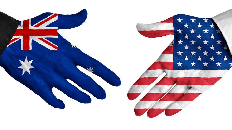 A stock image of a hand painted with an Australian flag reaching out to hand with an American flag