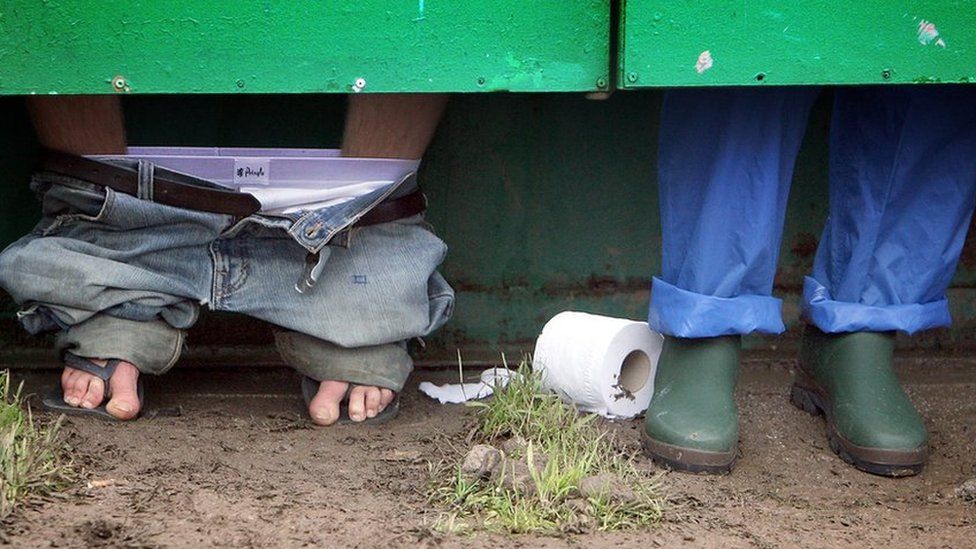 Festival goers (pictured with just their lower legs showing under a toilet door) use the toilets at Glastonbury Festival in Somerset, UK