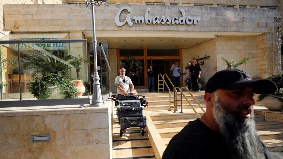A man pushing a cart carrying media equipment out of the Ambassador Hotel in Jerusalem while five people stand near the entrance