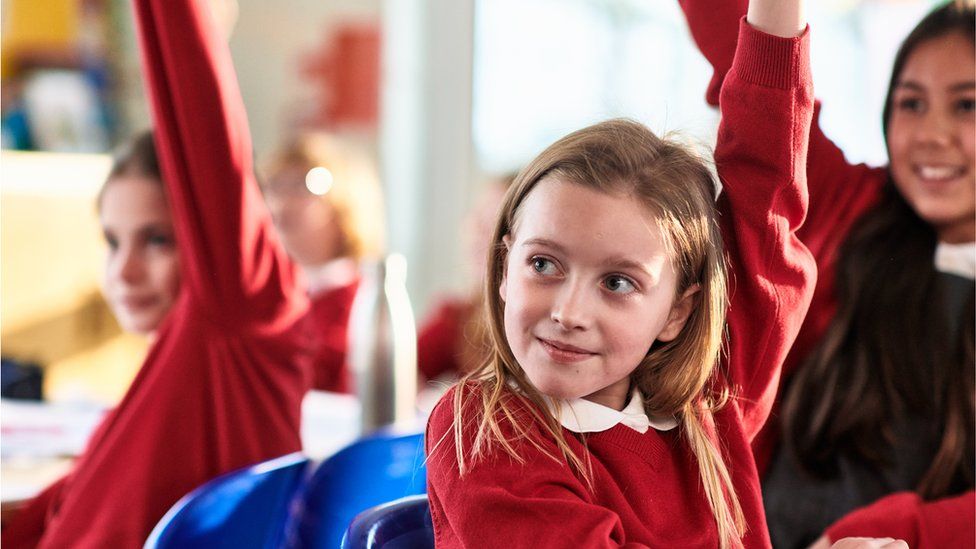 Primary school girl with blonde hair smiling with hand up in class