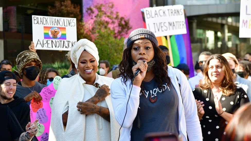 Выступает Блоссом С. Браун, а организатор митинга «Встаньте в солидарность» Эшли Мари Престон наблюдает за сотрудниками Trans и их союзниками на забастовке Netflix в знак протеста против специального мероприятия Дэйва Чаппеля 20 октября 2021 года в Лос-Анджелесе, Калифорния