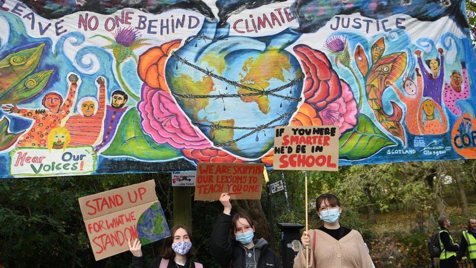 Colourful mural at COP26 in Glasgow saying 'leave no one behind climate justice'. Three young women in front of mural with protest signs.