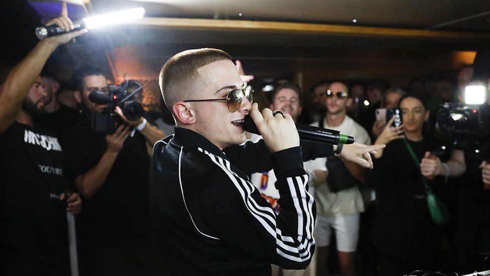 ArrDee holding a balck microphone to his lips, while there are multiple people in the back holding camera phones, lights and cameras recording him. His left hand is pointing at the crowd. He is wearing dark sunglasses, with the side of his visible. He is also wearing black adidas tracksuit top with the three white stripes down the side of his right arm.