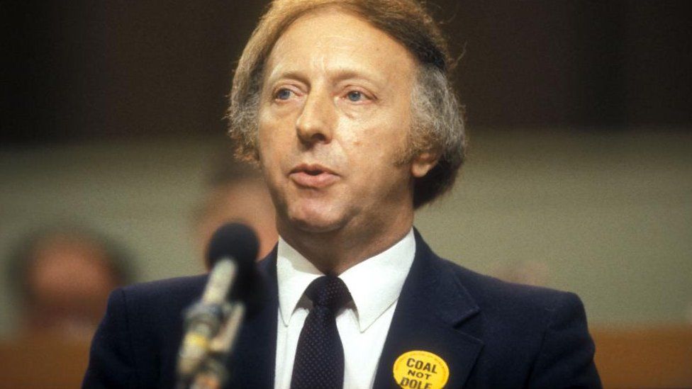 Arthur Scargill in front of microphone wearing "COAL NOT DOLE" sticker on lapel