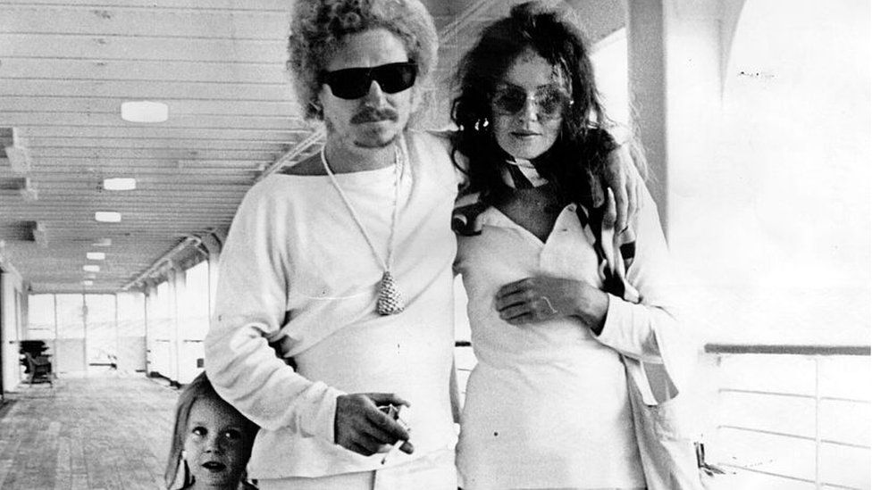 A black and white photo of a man and woman both wearing sunglasses indoors with a young girl next to them
