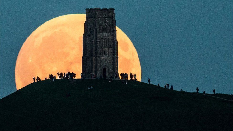 A huge red-tinged moon rises behind a hill and St Michael's Tower. People gathered by the tower.