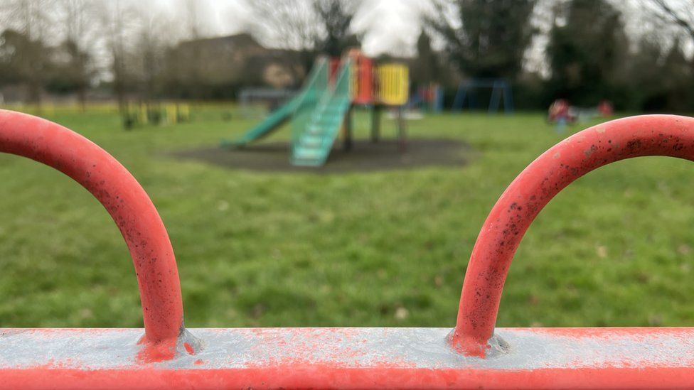 A children's play area. Metal railings in the foreground are visible, with swings and a slide in the background