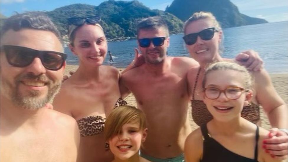 Four adults and two children posing for a selfie on a beach. The sun is shining and they all look happy.