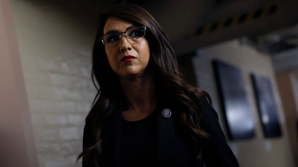 A photo taken form a low angle, Lauren Boebert looks to the right as she walks down a corridor