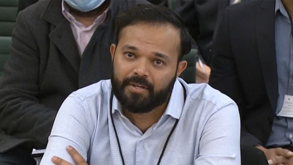 Azeem Rafiq, a bearded man at an official hearing. He is wearing a light blue shirt with a black lanyard around his neck and he looks emotional.