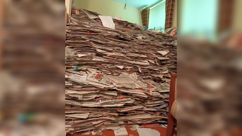 A room piled high with newspapers. The window is barely visible behind the pile