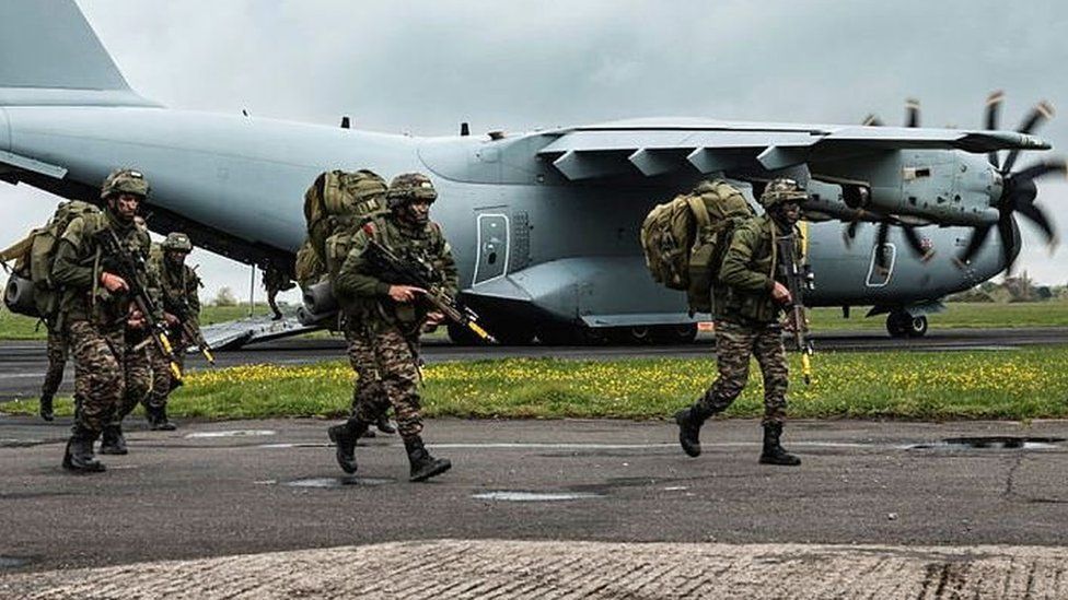 Military personnel in uniform, carrying guns, walking alongside a plane.