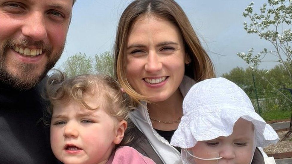 a mum, dad and two children the baby wearing a sunhat and with a nasal feeding tube