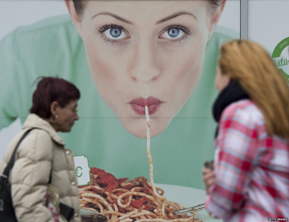 Women walk in front of a commercial featuring a woman eating pasta, outside a restaurant in Berlin