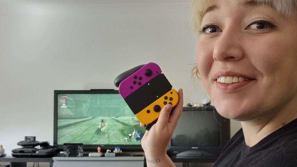Sky is at the right-hand side of the frame, smiling. She's holding up a Nintendo Switch controller, and in the background a TV, surrounded by different games consoles, is playing a Zelda game