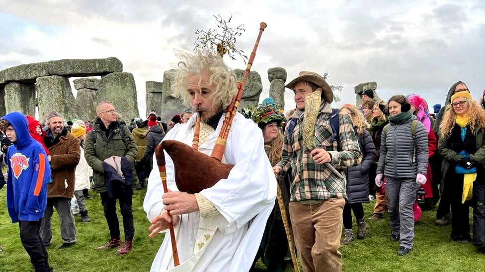 Crowds at Stonehenge including a man wearing a white robe while playing the bag pipes