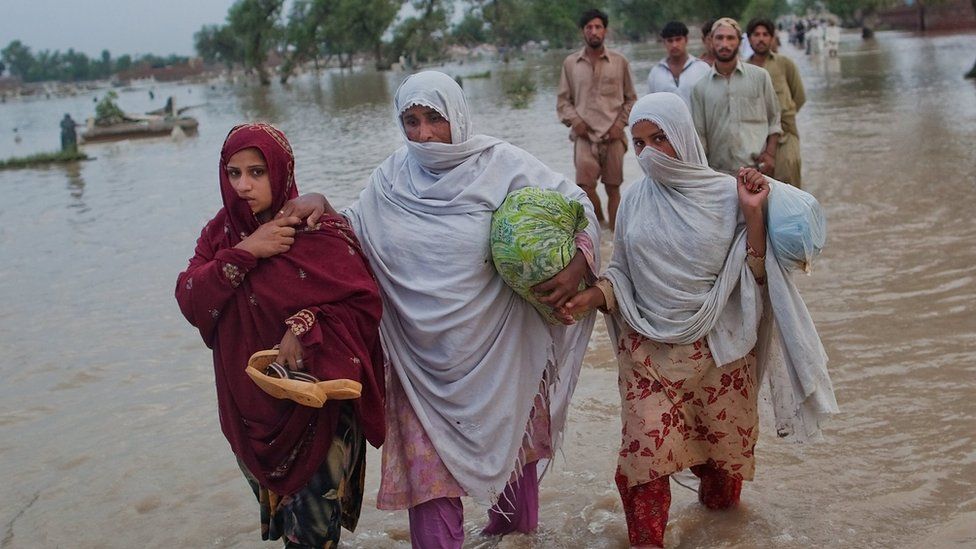 Three women in Pakistan wade through floodwater, carrying their belongings