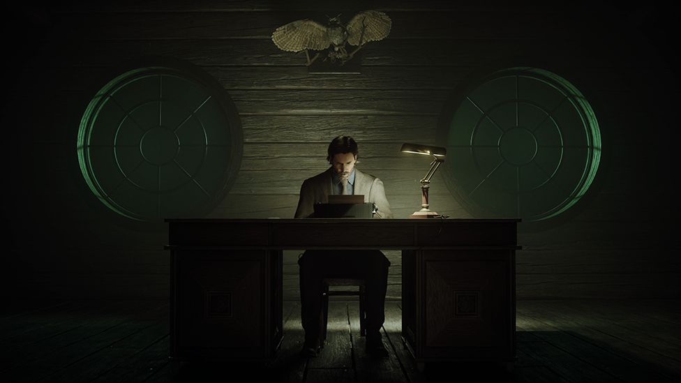 A man in a grey suit sits at a dimly lit desk, at a typewriter. The only source of light is an angle poise light to his left. He looks deep in concentration as he types away. We can just make out an eerie stuffed owl held on the wooden wall behind him by twigs. To either side of him, two identical circular objects that resemble ovesized celluloid film canisters appear to be balanced on the edge of the desk, lending a surreal element to the already sinister air of the image.