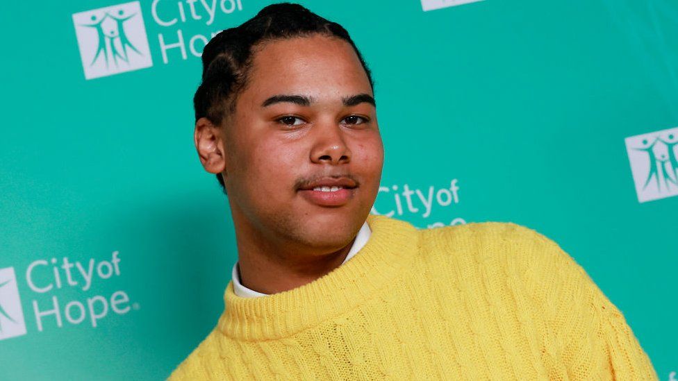 Singer songwriter Sekou Sylla pictured at an event in 2022. Sekou is a 19-year-old man with short black hair which he has braided. He has a small moustache and brown eyes and smiles slightly at the camera. He wears a yellow knitted jumper over a white shirt.