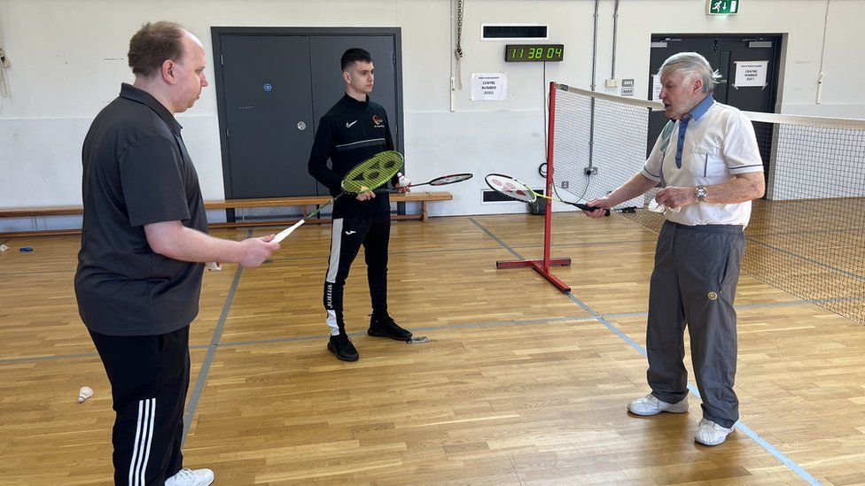 Colin Bedford holds out a badminton racket as he coaches two players on the court