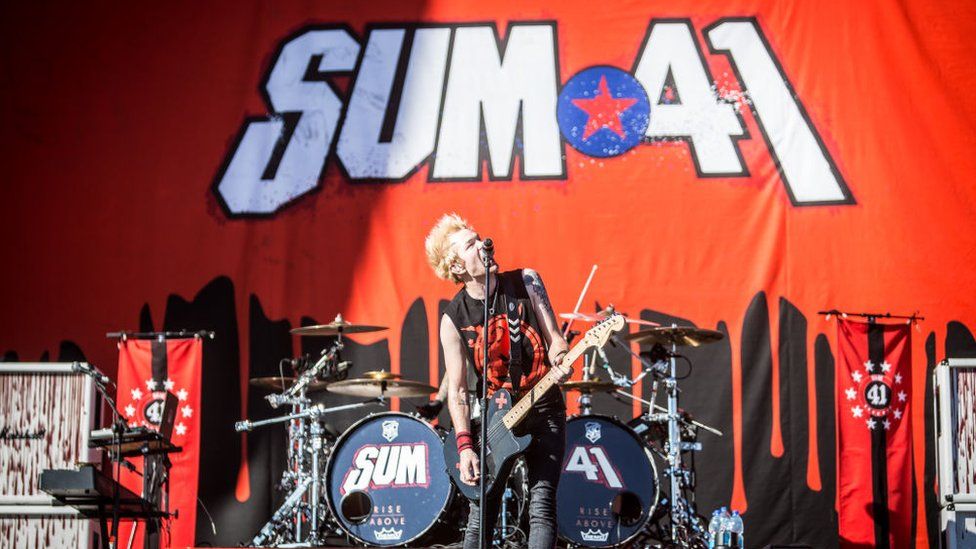Deryck Whibley of Sum 41 performing in Italy in 2017. Deryck is a white man in his 40s with bleached blonde hair. He wears a sleeveless black top with a red pattern on it. He plays a black guitar and stands in front of the band's drum kit. The staging is red with Sum 41's branding emblazoned on it in white font