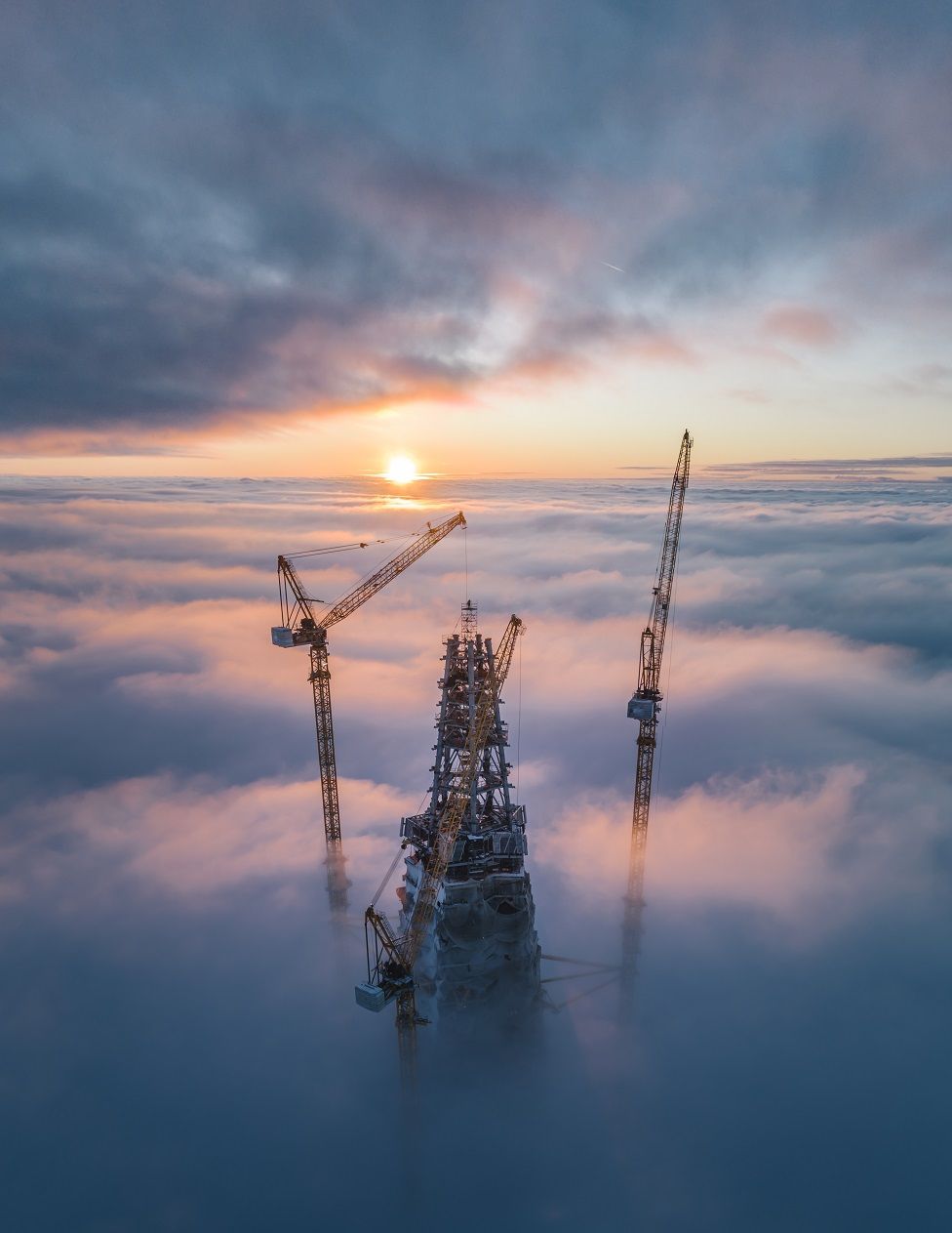 A skyscraper and two cranes poking above clouds