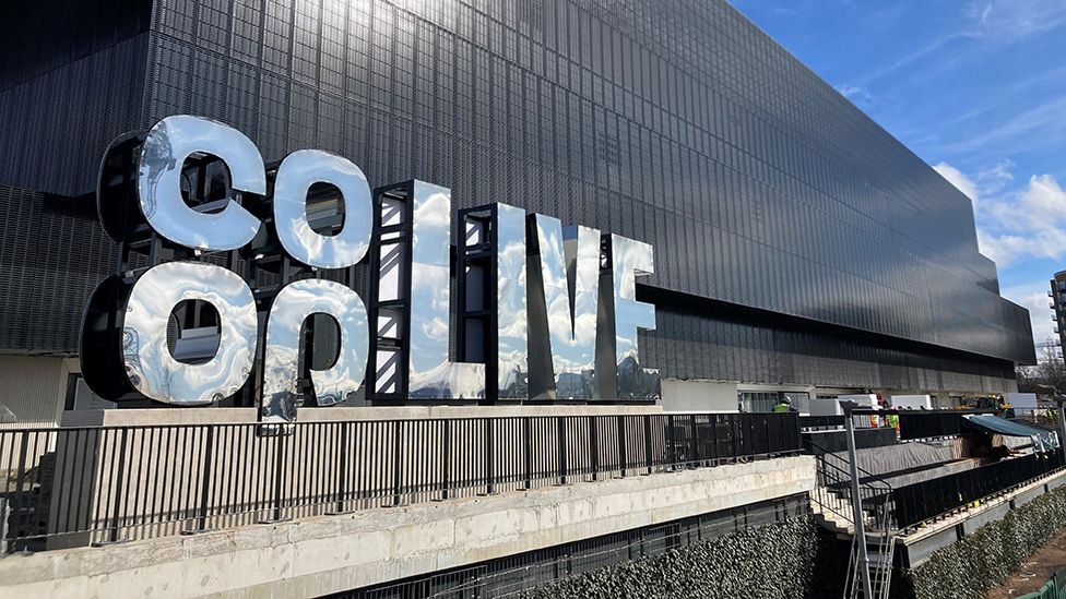 A large, black and windowless building stretches out behind a reflective silver sign spelling out the words "Co-Op Live". The sun is shining brightly and there are workers visible at the foot of the building, suggesting some construction work is under way.