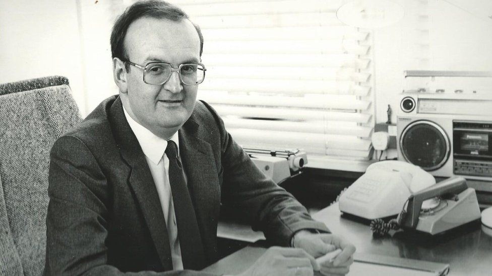 Black and white photo of Paul Deal in 1984, wearing glasses and sat at a desk beside two phones and a radio