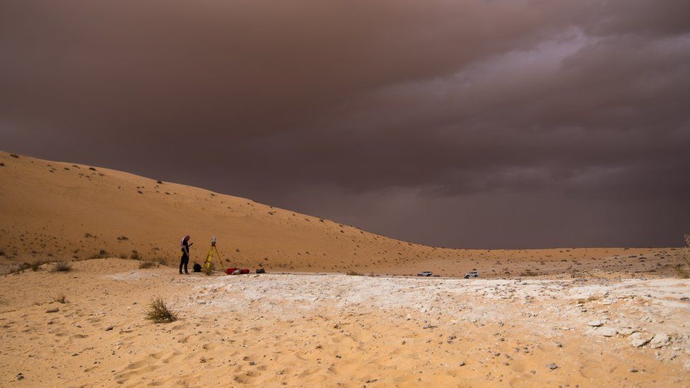 A bright desert against a dark, stormy looking sky. A scientist stands in middle distance.
