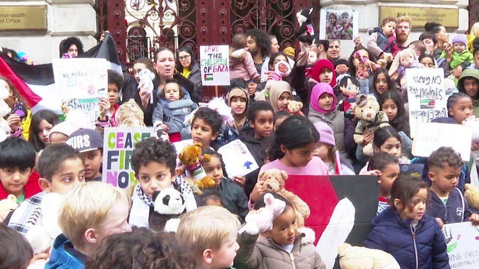 Dozens of children gathering with toys and placards in front of the gates of the Foreign Office