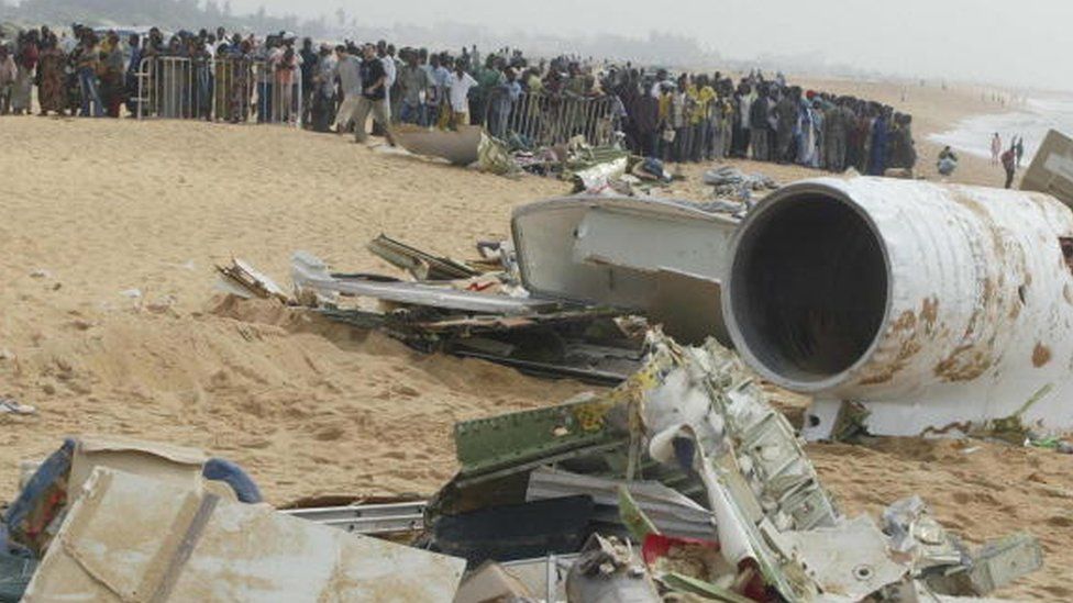 人们站在障碍物后面看着贝宁坠毁的飞机残骸（2003 年 12 月 26 日）