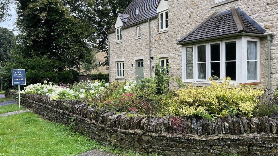 A traditional Cotswold stone house with a 'for sale' sign outside