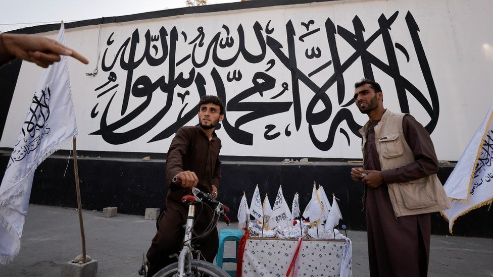 Men react while they sell Taliban flags of the Islamic Emirate of Afghanistan in front of a mural with the same flag, in front of the former U.S. embassy in Kabul, Afghanistan October 8, 2021.