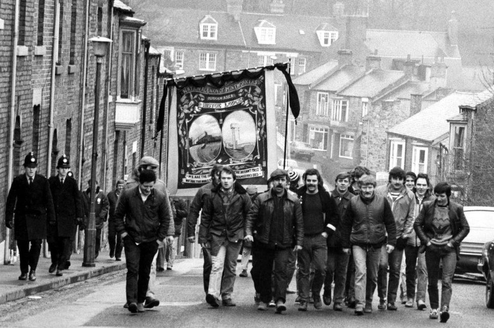 Black and white photo from 1984 showing a large group of men walking up a street in Durham with a banner and police officers off to one side