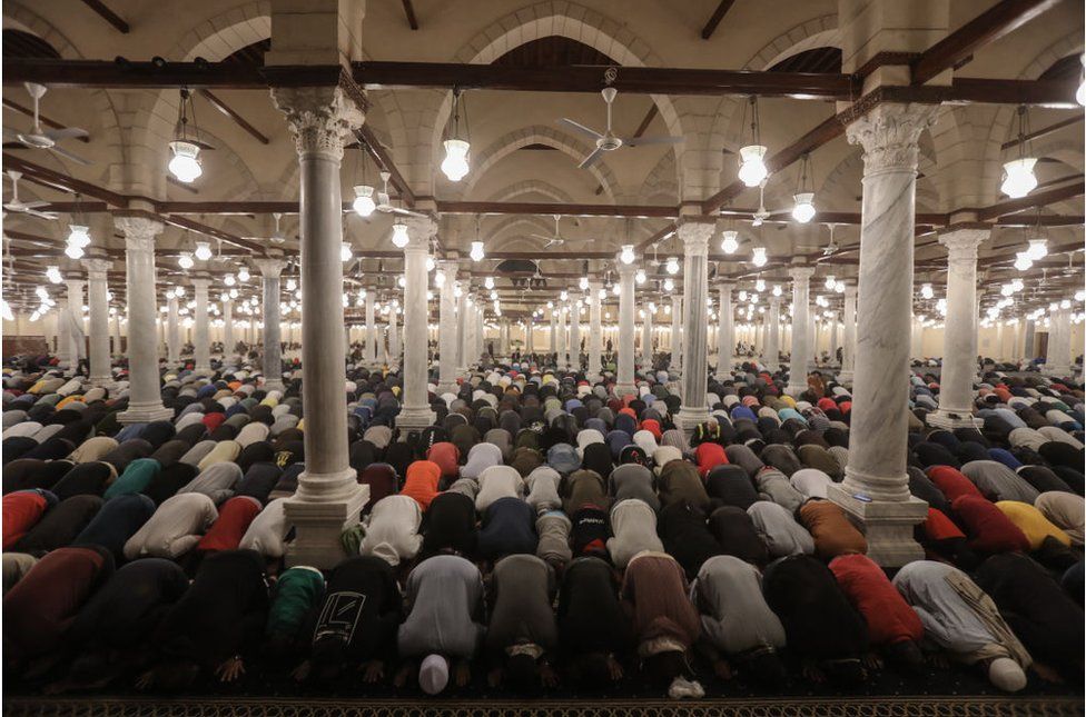 People praying in a mosque. They are all wearing different colour clothes and are bowing down.