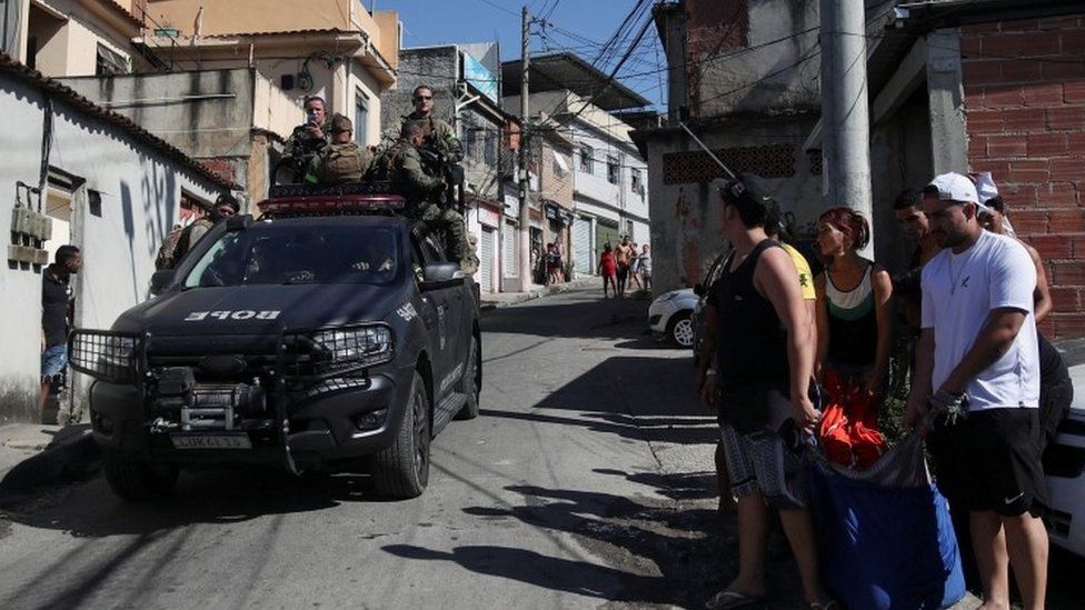 Police officers drive past people carrying a dead body during a police operation against drug gangs in Alemao