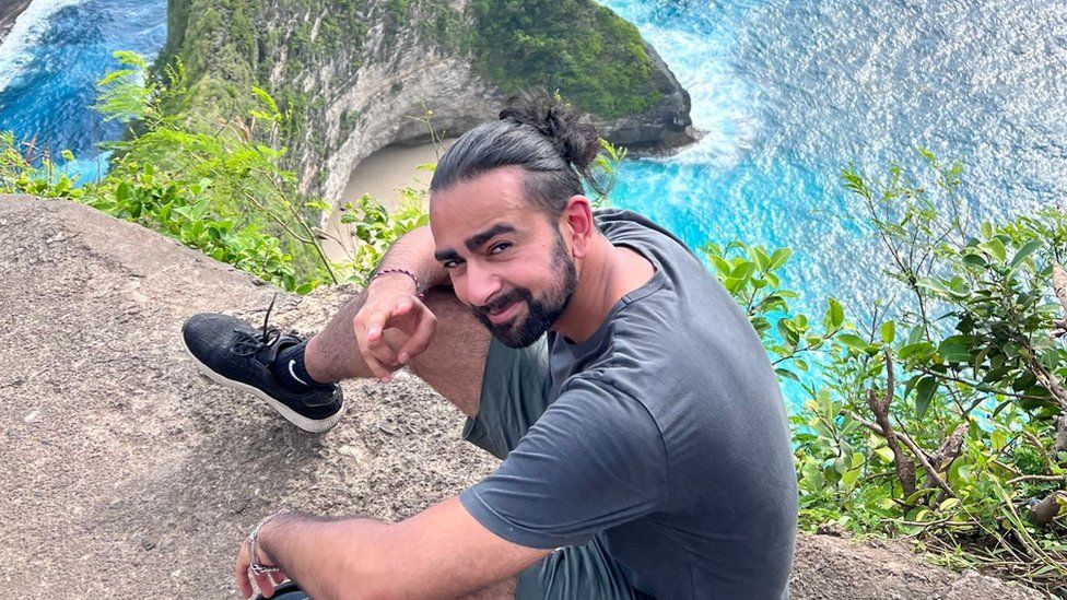 Aiz Hussain smiling for the camera, which is above him, as he sits on the edge of a rock at the top of a cliff. His dark hair is pulled back into a ponytail and he has a dark beard. He is wearing a blue/grey T-shirt and and similar colour shorts, with black socks and black trainers on. He is making the peace sign with his right hand towards the camera. Behind him you can see the bright blue sea at the bottom of the cliff and a small bit of a beach at the bottom of a rocky outcrop