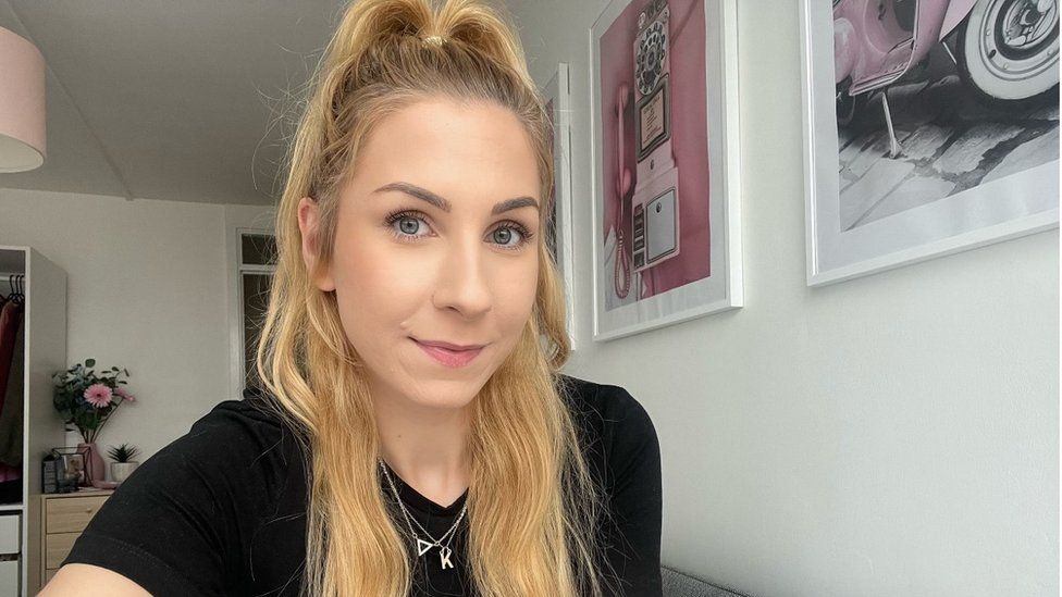Kat Leech, a 27-year-old white woman with long blonde hair tied up in a half-up-half-down style. She wears a black T-shirt and a small gold necklace. She has blue eyes and smiles at the camera as she takes a selfie in her bedroom. Behind her are pink and grey prints on the white wall and a vase of pink flowers on a bedside cabinet.