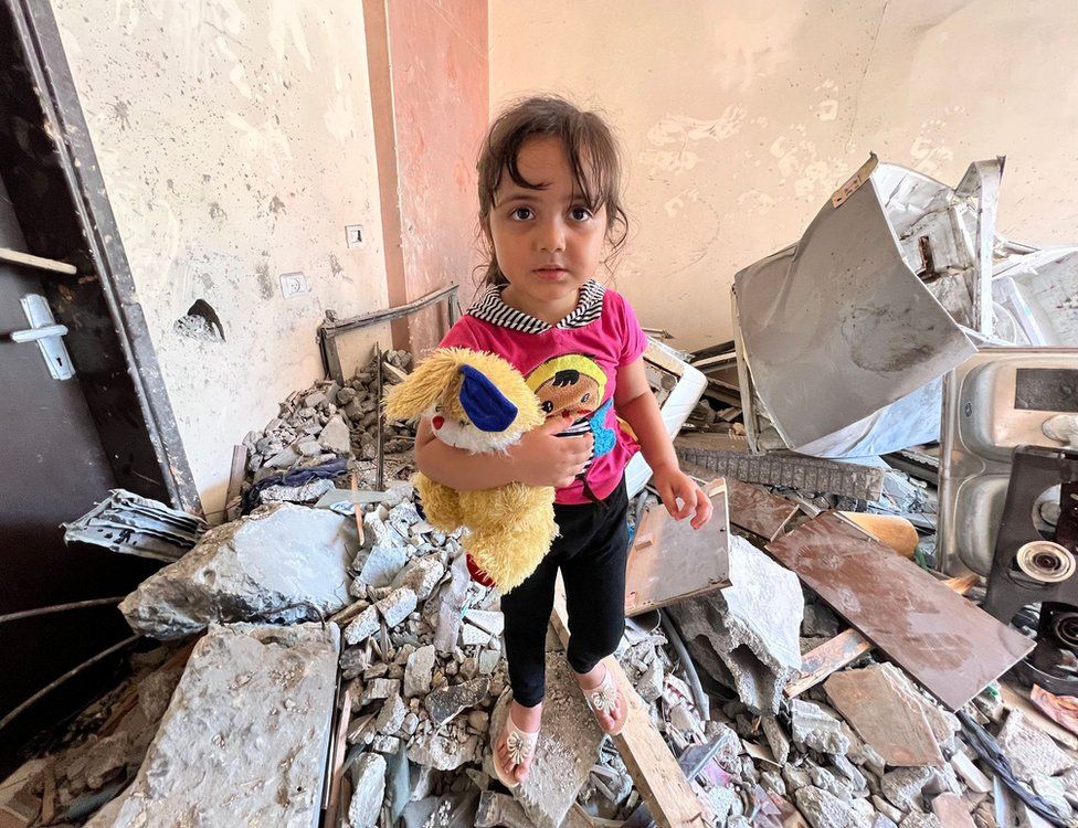 Tuta standing on rubble of a destroyed home carrying a yellow teddy bear