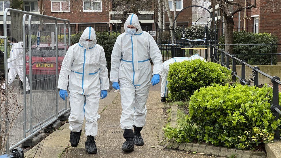 People in white forensics suits walking outside some flats.
