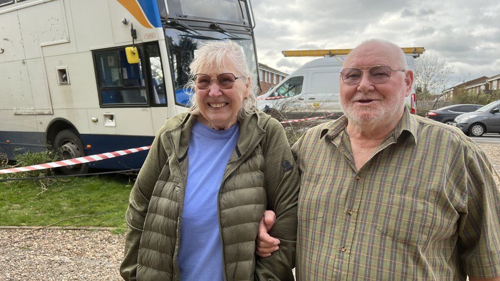 Mr and Mrs Bull linking arms and smiling stood by the bus that crashed into their front garden