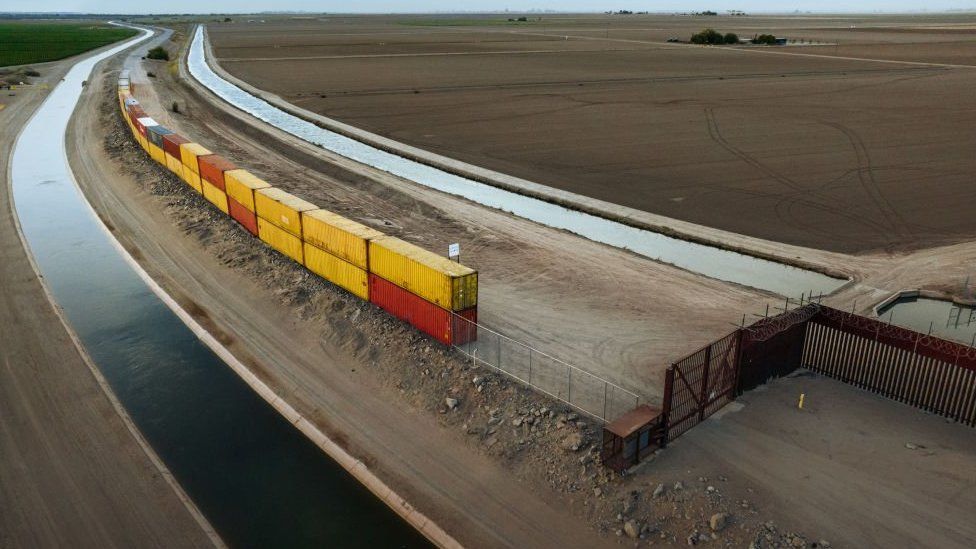 Фотография желтых и красных грузовых контейнеров, сложенных попарно, чтобы заполнить пробел в недостроенной стене на границе США и Мексики