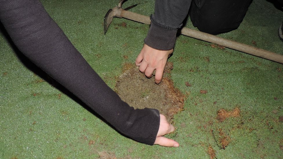 Two hands reaching down to fill golf course hole with cement. Pick-axe seen in background