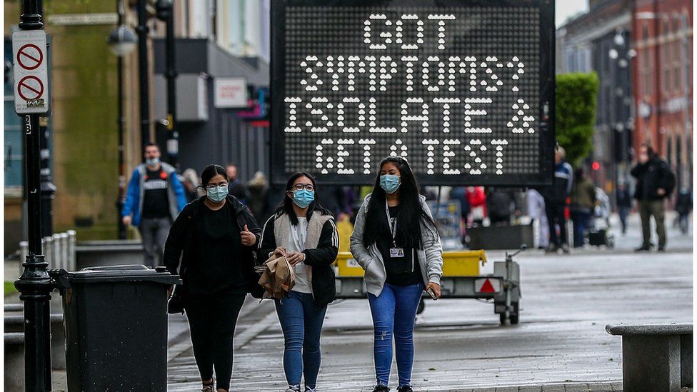 Three women in face masks walking in Bolton with an electronic sign telling people with symptoms to isolate
