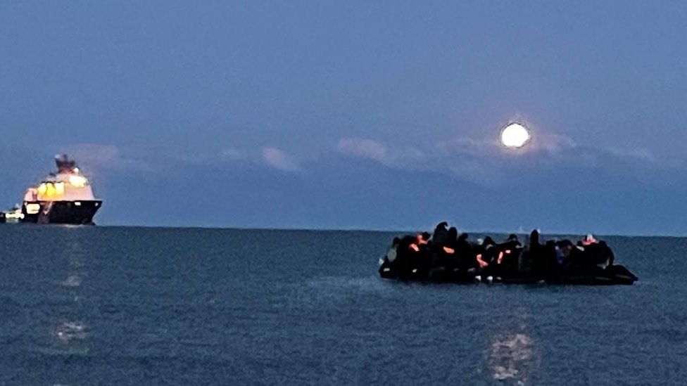 An overcrowded inflatable boat heads out to sea in the English Channel under moonlight. A French coastguard ship is on the horizon.