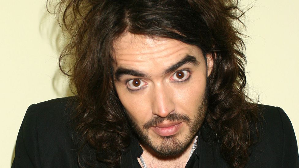 Portrait of Russell Brand looking at the camera. He has facial hair and long hair